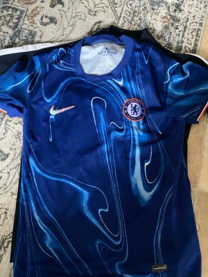 Chelsea FC blå matchtröja Nike - Säljer en blå Chelsea FC matchtröja från Nike med unikt mönster i olika blå nyanser. Tröjan har korta ärmar, rund hals och Chelsea-logga på bröstet. Perfekt för fotbollsträning eller att bära som supporter. Materialet är lätt och andas.