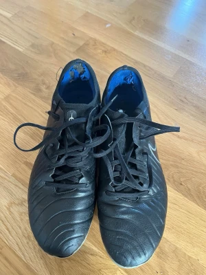 Svarta Nike tiempo fotbollsskor med snörning - Säljer ett par svarta Nike tiempo legend 10 PRO fotbollsskor med klassisk snörning och diskret Swoosh på sidan. Skorna har en stilren design med struktur på ovandelen och blå insida. Perfekta för dig som vill ha bra grepp och kontroll på planen. Insidan trasig men annars bra skick 