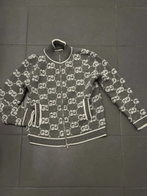 Grå stickad kofta från Gucci - Snygg grå stickad kofta från Gucci med hel dragkedja och vit logotypmönster över hela plagget. Ribbstickade muddar och krage med vita detaljer. Perfekt för dig som gillar exklusiv streetstyle och vill sticka ut med ett lyxigt plagg. Pris kan diskuteras 