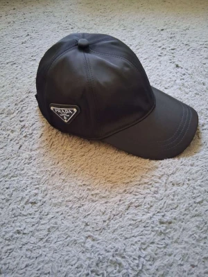 Svart keps från Prada i nylon - Snygg svart keps från Prada i glansigt nylonmaterial med klassisk böjd skärm och ikonisk trekantslogga på sidan. Perfekt accessoar för en trendig och sportig look.