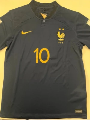 Sprillans Ny #10 Mbappe Frankrike Matchtröja Nike  - Mörkblå fotbollströja från Nike med Frankrikes landslagslogga, nummer 10 och Mbappé på ryggen. Tröjan har gula detaljer och två stjärnor ovanför tuppen. Perfekt för dig som älskar fotboll och vill stötta Les Bleus.