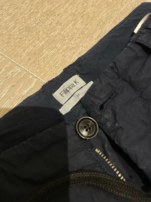 Mörkblå kostymbyxor Filippa K - Snygga mörkblå kostymbyxor från Filippa K med klassisk passform och knappgylf. Byxorna har raka ben och är perfekta för en stilren look. Materialet känns lätt och bekvämt, och färgen är djupblå vilket gör dem enkla att matcha.