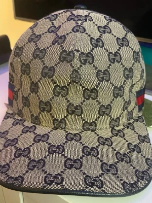Gucci keps med GG-mönster - Säljer en beige och marinblå keps från Gucci med det klassiska GG-monogrammönstret över hela kepsen. På sidan finns en ikonisk röd och marinblå rand. Kepsen har böjd skärm och justerbar rem bak. Materialet är canvasliknande tyg.