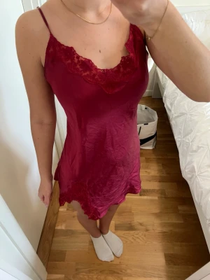 Vinrött nattlinne med spetsdetaljer - Säljer ett vinrött nattlinne med tunna axelband och fina spetsdetaljer vid urringningen och nederkanten. Materialet är glansigt och mjukt, vilket ger en lyxig känsla. Perfekt för dig som vill ha något bekvämt och snyggt att sova i.