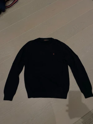 Mörkblå stickad tröja från Morris - Snygg mörkblå stickad tröja från Morris med rund halsringning och diskret röd logga broderad på bröstet. Tröjan har ribbade muddar vid ärmslut och nederkant, perfekt för en stilren och enkel look.