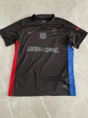 Svart FC Barcelona x Cactus Jack tröja - Säljer en svart FC Barcelona x Cactus Jack t-shirt från Nike. Tröjan har klubbmärke på bröstet, röd Nike-logga, grafiskt tryck med 'Cactus Jack' och röda samt blå paneler på sidorna. Perfekt för dig som gillar unika fotbollströjor och streetwear.