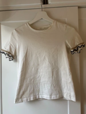Vit t-shirt med volangkant - Säljer en vit t-shirt från reserved med korta ärmar och volangkant i svart och guld. Ärmsluten har en snygg virkad detalj som ger tröjan en unik touch. Perfekt för dig som vill ha något enkelt men ändå speciellt. Storleken är S💞