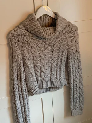 Grå croppad polotröja med flätmönster - Säljer en grå, croppad stickad polotröja med grovt flätmönster både fram och på ärmarna. Tröjan har bred polokrage och ribbade muddar. Perfekt för dig som gillar en mysig och trendig look till hösten eller vintern.                                                                                      Har tyvärr inte kvar lappen, så det är oklart vart den är ifrån, samt storlek, men skulle gissa H&M. 