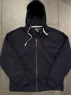 Marinblå hoodie från Polo Ralph Lauren - Snygg marinblå hoodie med dragkedja från Polo Ralph Lauren. Klassisk design med vit dragsko i huvan och röd broderad logga på bröstet. Tillverkad i bomull och polyester för en skön och avslappnad känsla (Se bilder för mått).