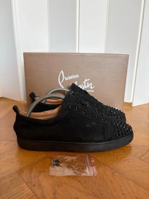 Svarta Christian Louboutin sneakers med spikar  - Säljer ett par svarta sneakers från Christian Louboutin i mocka med svarta nitar på tån. Skorna har en låg profil, svarta skosnören och en klassisk siluett. Perfekta för dig som vill sticka ut med en edgy och lyxig stil. Dem har tyvär en skavank, längst bak på skon så är de limmade syns på bild. Och så är de lite slitna i hälen. Skolåda och extra spikar medföljer såklart. Tveka inte att fråga om ni har några frågor😀