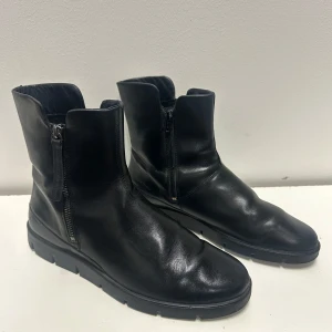 Svarta boots med dragkedja från Ecco 41 - Snygga svarta boots från Ecco i slätt läder med dragkedja på sidan och robust sula. Klassisk design med rundad tå och diskret söm längs kanten. Perfekta för dig som gillar stilrena och tidlösa kängor.