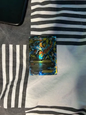 Pokémonkort M Charizard EX - Säljer ett samlarkort från Pokémon, M Charizard EX. Kortet har en färgstark och glansig design med blå, gul och svart färg. Det är ett EX-kort med 230 HP och attacken Wild Blaze. Perfekt för dig som gillar Pokémon och samlarkort.  