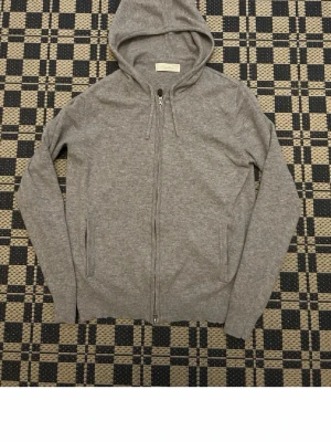 Grå hoodie med dragkedja - Snygg grå hoodie från sayless med dragkedja framtill och två fickor. Tillverkad i mjukt material och har ribbade muddar vid ärmslut och nederkant. Klassisk huva med dragsko för extra komfort. Cashmire 