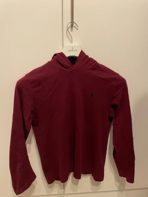 Vinröd hoodie från Ralph Lauren - Snygg vinröd hoodie från Ralph Lauren med klassisk huva och diskret logga på bröstet. Tillverkad i mjukt material och har långa ärmar. Perfekt för en avslappnad och stilren look. Passar även vissa med storlek d