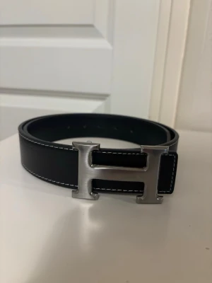 Svart bälte med H spänne - Snyggt svart bälte med ett stort, silverfärgat H-spänne. Bältet har vita kontrastsömmar längs kanterna och är gjort i slätt skinn. Perfekt accessoar för att lyfta vilken outfit som helst. Perfekt skick i storlek 100 helt oavänt