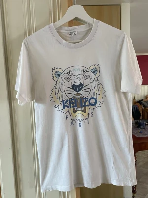 Vit Kenzo t-shirt med tigertryck - Säljer en vit t-shirt från Kenzo med det ikoniska tigertrycket och Kenzo Paris-loggan framtill. T-shirten har rund hals, korta ärmar och är gjord i mjuk bomull. Perfekt för dig som vill ha en stilren men ändå statement look. Herr! Stl M  litet hål. Annars fint skick! 