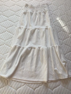 Vit långkjol med volanger - Superfin vit långkjol med volanger och resår i midjan. Kjolen har en luftig och flowy känsla, perfekt för varma dagar. Tillverkad i ett mjukt tyg som känns skönt mot huden. Enkel att matcha med olika toppar för en avslappnad look.