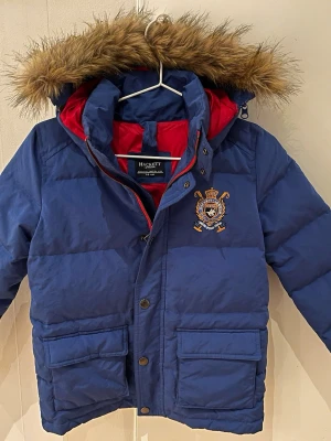 Blå dunjacka från Hackett London, barn - Blå dunjacka från Hackett London för barn. Storlek 122/128, ålder 7-8 år. Jackan har en röd insida, avtagbar huva med fejkpäls och broderad patch på bröstet. Jackan har stora fickor, dragkedja och knappar framtill samt en röd siffra 1 på ärmen. Perfekt för kalla dagar! Yttertyg 61% polyester och 39% nylon. Fyllning anddun. Maskintvätt 30 grader. 