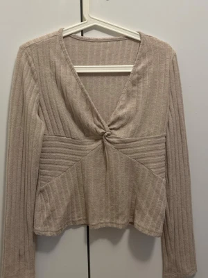 Beige ribbad topp med knut - Trendig beige långärmad topp med ribbad struktur och snygg knutdetalj framtill. Djup v-ringning och figurnära passform ger en modern look. Perfekt att matcha med jeans eller kjol för en cool stil.