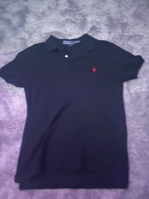 Svart pikétröja från Polo Ralph Lauren - Klassisk svart pikétröja från Polo Ralph Lauren med röd broderad logga på bröstet. Slim fit-modell med korta ärmar och krage med knappar. Perfekt för en stilren och enkel look.