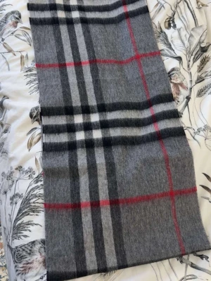 Berberry halsduk  - Snygg grå halsduk med klassiskt rutigt mönster i svart, vitt och rött. Halsduken är rektangulär och har en mjuk känsla, perfekt att svepa runt halsen för extra stilpoäng. Passar till de flesta outfits och ger en tidlös vibe.