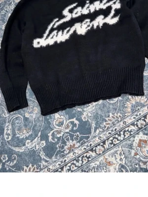 Svart stickad Saint Laurent-tröja - Cool svart stickad tröja från Saint Laurent med vit logotyp i grafisk stil över bröstet. Tröjan har långa ärmar och bred ribbstickning vid ärmslut och nederkant. Perfekt för dig som gillar streetwear och designerplagg.