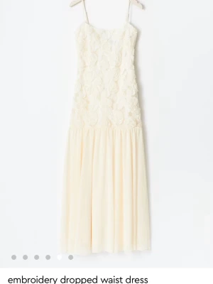 Ljus långklänning med broderade blommor - Superfin ljus långklänning med tunna axelband och broderade blommor upptill. Klänningen har en dropped waist och ett flowy fall. Perfekt för sommarens alla tillfällen!