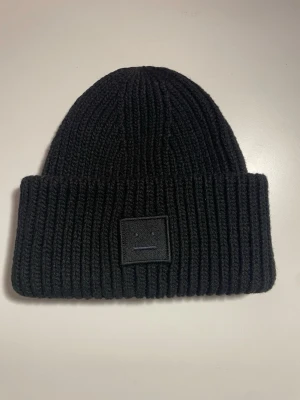 Svart mössa från Acne Studios - Ribbstickad svart mössa från Acne Studios med uppvikt kant och det klassiska ansiktsloggan framtill. Tillverkad i mjuk ull som håller dig varm och stilren under kalla dagar. Perfekt accessoar för en minimalistisk look.