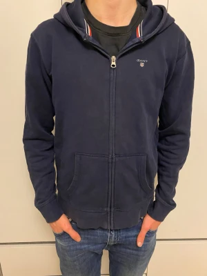 Mörkblå hoodie från GANT - Snygg mörkblå zip up tröja från gant som jag säljer för att jag behöver pengar och vill rensa gamla kläder. Varan är köpt för 500kr. Priset kan diskuteras och hör gärna av dig om du är sugen på att köpa 