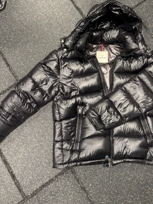 Svart dunjacka från Moncler - Snygg svart dunjacka från Moncler helt ny med glansig finish och huva. Jackan har två dragkedjefickor framtill och klassisk Moncler-logga på insidan. Perfekt för kalla vinterdagar och riktigt trendig look.