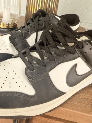 Nike Dunk Low svart/vit sneakers - Säljer ett par Nike Dunk Low sneakers i svart och vitt läder. Klassisk siluett med svart swoosh på sidan, svarta skosnören och vit tåbox med perforeringar. Yttersulan är svart och mellansulan vit. Perfekta för dig som gillar streetwear och tidlös stil.