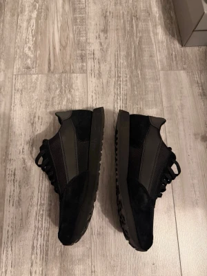 Svarta sneakers från Armani Exchange - Snygga svarta sneakers från Armani Exchange med diskret logga på plösen. Skorna har en mix av mocka och meshmaterial, samt svarta snören och sula med grovt mönster. Perfekta för en stilren och modern look.