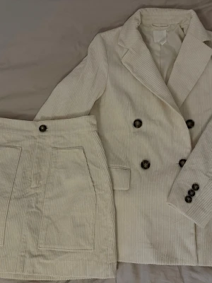 Vit manchesterkostym med kjol - Snygg vit manchesterkostym med dubbelknäppt kavaj och matchande kjol. Kavajen har breda slag, dekorativa knappar och fickor. Kjolen är rak med stora fickor fram och knapp i midjan. Perfekt för dig som vill sticka ut med en retroinspirerad look. Kavaj strl: 32 (Passar även 34). Kjol strl: 34. Observera en liten fläck på vänster ärm som bör gå bort i tvätt!