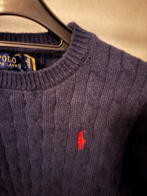 Mörkblå kabelstickad tröja Polo Ralph Lauren - Kabelstickad mörkblå tröja från Polo Ralph Lauren med rund halsringning och röd broderad logga på bröstet. Klassisk och tidlös design i mjukt material, perfekt för lager-på-lager-stil.