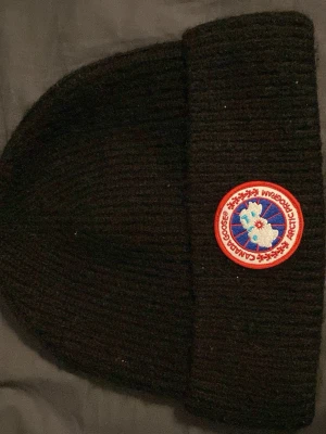 Svart mössa från Canada Goose - Säljer en svart ribbstickad mössa från Canada Goose med klassisk rund logotyp framtill i rött, vitt och blått. Mössan har uppvikt kant och är tillverkad i ett mjukt, värmande material som passar perfekt för kalla dagar.