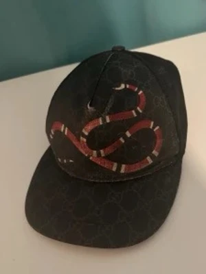 Gucci keps med ormtryck - Snygg svart keps från Gucci med diskret GG-mönster och ett stort rött och vitt ormtryck på framsidan. Klassisk böjd skärm och justerbar baksida. Perfekt för dig som vill sticka ut med en exklusiv accessoar.