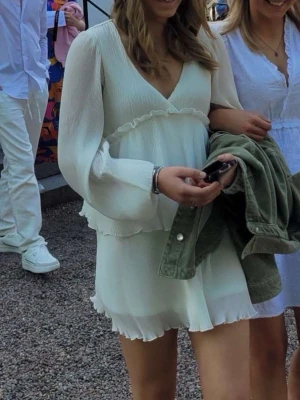 Vit kort omlottklänning med volang - Söt vit kortklänning med omlott-design och volangdetaljer. Klänningen har långa, lätt transparenta ärmar och v-ringad hals. Tillverkad i ett tunt, luftigt material som ger en flowy känsla. Perfekt för sommarens alla tillfällen. Från Nelly 