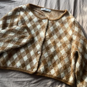 Kofta från Zara - Säljer en mysig rutig kofta från Zara i brunt och vitt. Koftan har en klassisk design med knappar framtill och långa ärmar. Perfekt för att hålla sig varm under kyligare dagar. Jättefin till layering
