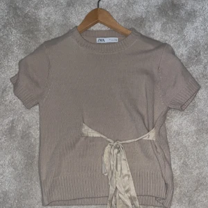 Beige kortärmad tröja från Zara - Säljer en stilren beige kortärmad tröja från Zara. Tröjan har en knytning i midjan och ribbade kanter vid ärmslut och nederkant. Perfekt för en avslappnad look. Priset går att diskutera