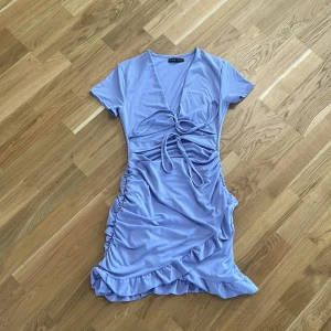 Ljuslila omlottklänning - Snygg ljuslila omlottklänning med volangdetaljer och knytning framtill. Klänningen har korta ärmar och en v-ringad design som ger en trendig look. Perfekt för sommarens alla tillfällen!