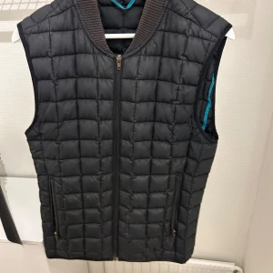 Svart quiltad väst med dragkedja - Snygg svart quiltad väst med dragkedja framtill och ribbad krage. Perfekt för lager-på-lager under kyligare dagar. Två praktiska fickor med dragkedja på sidorna.