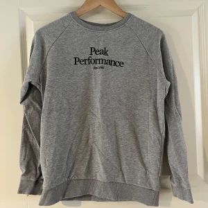 Grå tröja från Peak Performance - Säljer en stilren grå tröja från Peak Performance med svart text på bröstet. Tröjan har långa ärmar och ribbade muddar vid ärmslut och nederkant. Kontakta ifall frågor