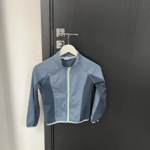 Blå vindjacka med dragkedja - Snygg blå vindjacka med ljusgrön dragkedja. Perfekt för blåsiga dagar. Jackan har en hög krage och långa ärmar för extra skydd.