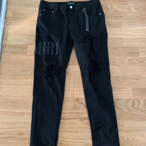 Svarta jeansbyxor från Amiri - Snygga svarta jeansbyxor från Amiri med slitna detaljer och stort tryck på ena benet. Byxorna har en knappgylf och klassisk femficksdesign. Perfekta för en trendig look.