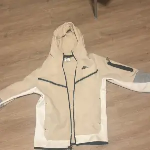 Snygg beige hoodie från Nike med grå detaljer och dragkedja. Den har en huva och långa ärmar med en unik ficka på ena ärmen. Perfekt för en sportig look eller avslappnad stil. Storlek herr xs köpt på jd sports 