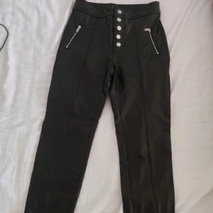 Svarta skinnbyxor med dragkedjor - Snygga svarta skinnbyxor med hög midja och knappar framtill. Byxorna har dragkedjor vid fickorna och en tight passform. Perfekta för en trendig look.