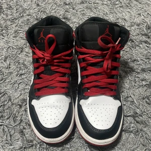 Nike Air Jordans i svart, vitt och rött - Snygga Nike Air Jordans i en klassisk färgkombination av svart, vitt och rött. Skorna har en hög design med röda snören och den ikoniska swoosh-loggan på sidan. Perfekta för dig som vill ha en stilren och sportig look.