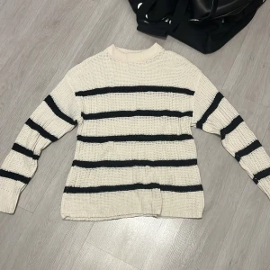 Randig stickad tröja - Säljer en mysig stickad tröja i beige med svarta ränder. Tröjan har en klassisk design med långa ärmar och rund hals. Perfekt för kyligare dagar.
