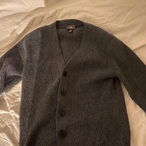 Grå kofta i ull - Säljer en stilren grå kofta från H&M i 100% ull. Koftan har en v-ringad design med knappar framtill och långa ärmar. Perfekt för kyligare dagar och ger en klassisk look. Passar bra till både jeans och kostymer.