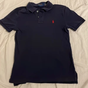 Mörkblå pikétröja från Ralph Lauren - Säljer en klassisk mörkblå pikétröja från Ralph Lauren med den ikoniska röda loggan broderad på bröstet. Tröjan har en stilren krage med knappar. Perfekt för en avslappnad men stilfull look. Passar både till jeans och chinos. Storleken är 14-16 men passar xs
